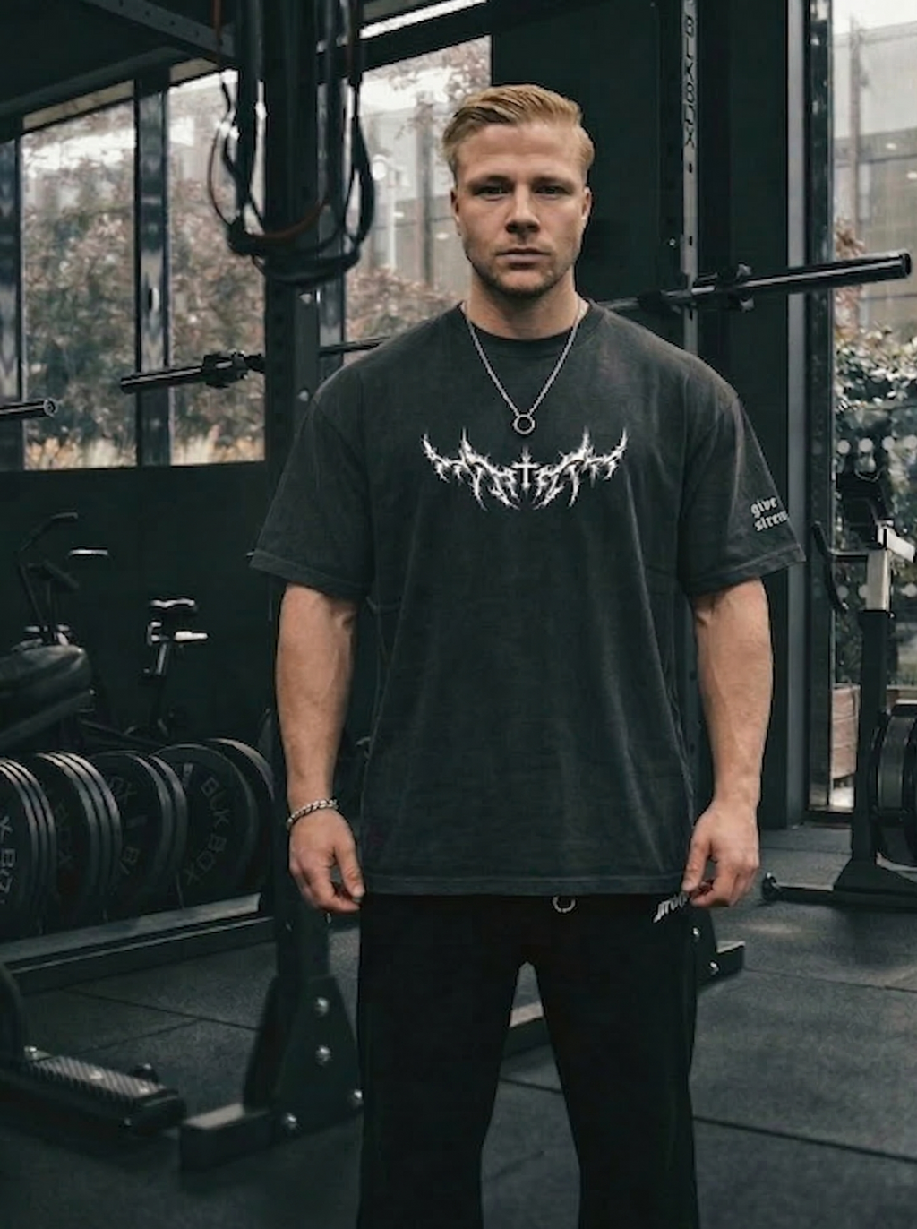 En man i ett gym som bär en svart oversized Tshirt där det är ett stort kristet kors i tribal likande mönster på bröstet och en text på armen där det står "give me strength" i en gotisk typografi.