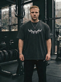 En man i ett gym som bär en svart oversized Tshirt där det är ett stort kristet kors i tribal likande mönster på bröstet och en text på armen där det står "give me strength" i en gotisk typografi.