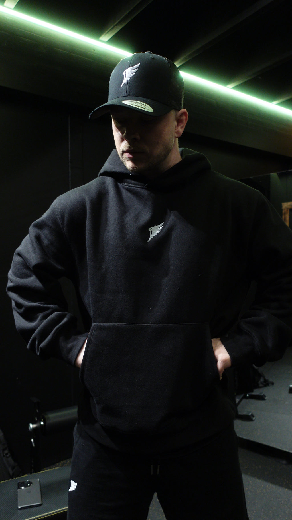 En man i ett gym som bär en svart keps och en svart oversized hoodie med en ving-logga broderad i grått på bröstet.