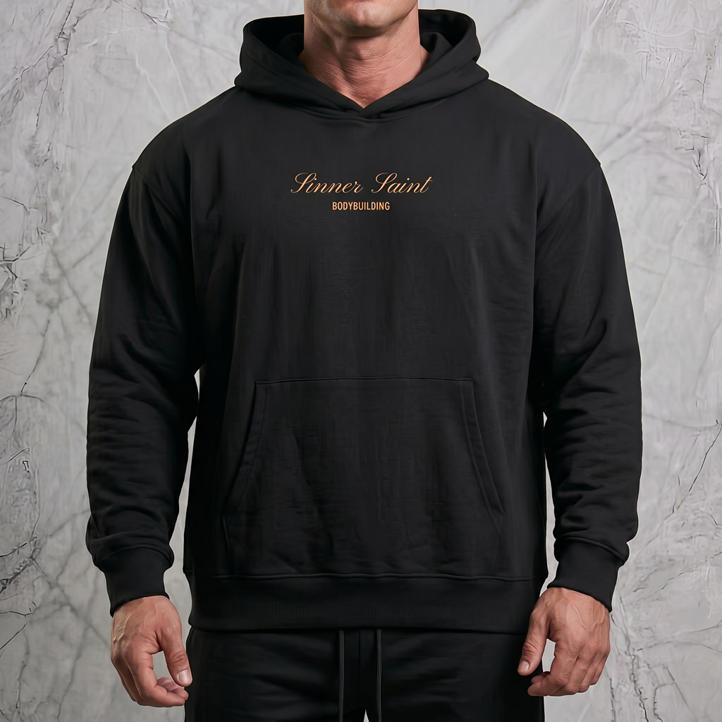 Man i en lyxig fotostudio som bär en svart herr och dam oversized hoodie med framficka och huva med en orange text på bröstet där det står "Sinner Saint Bodybuilding".
