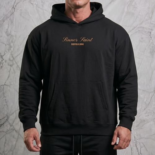 Man i en lyxig fotostudio som bär en svart herr och dam oversized hoodie med framficka och huva med en orange text på bröstet där det står "Sinner Saint Bodybuilding".