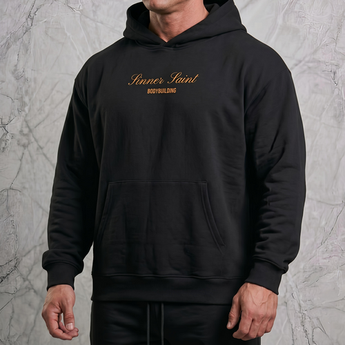 Man i en lyxig fotostudio som bär en svart herr och dam oversized hoodie med framficka och huva med en orange text på bröstet där det står "Sinner Saint Bodybuilding".