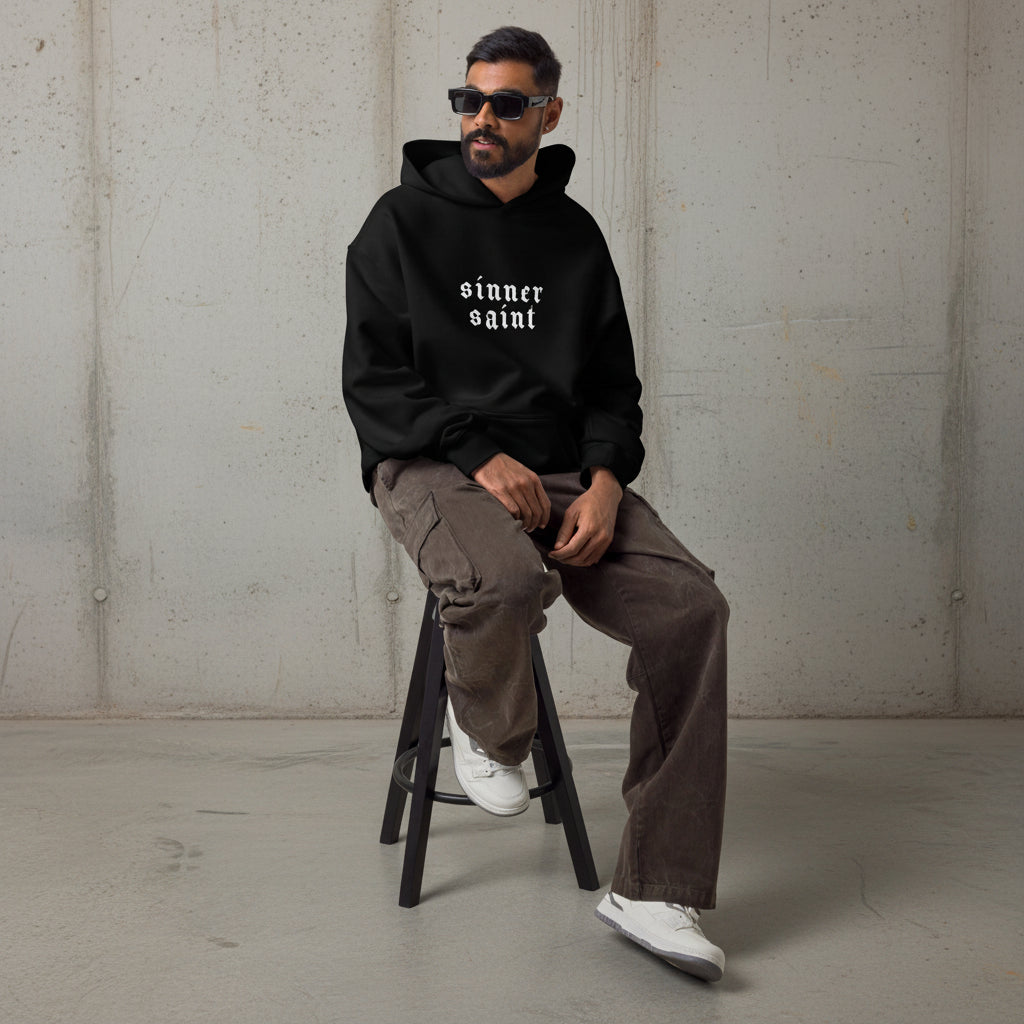 Man in black oversized activewear hoodie sitting on a stool with text on the chest saying "Sinner Saint".