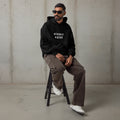 Man in black oversized activewear hoodie sitting on a stool with text on the chest saying "Sinner Saint".