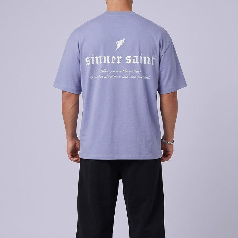 En man bakifrån i en fotostudio som bär en violett oversized tshirt där det står Sinner Saint, "When you feel like quitting, remember all of those who said you'd fail".